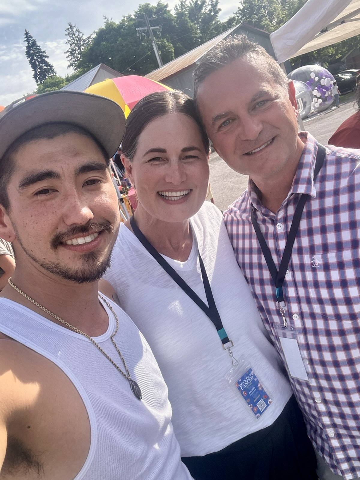 In this image, Jack (left), Jodie (middle), and Kevin (right) are smiling together at the Rock Out Cancer concert. Jack is wearing a gray cap and white tank top, Jodie has her hair pulled back and is in a white shirt, and Kevin is in a checkered shirt. They’re enjoying the sunny day at the event, with tents and people visible in the background.